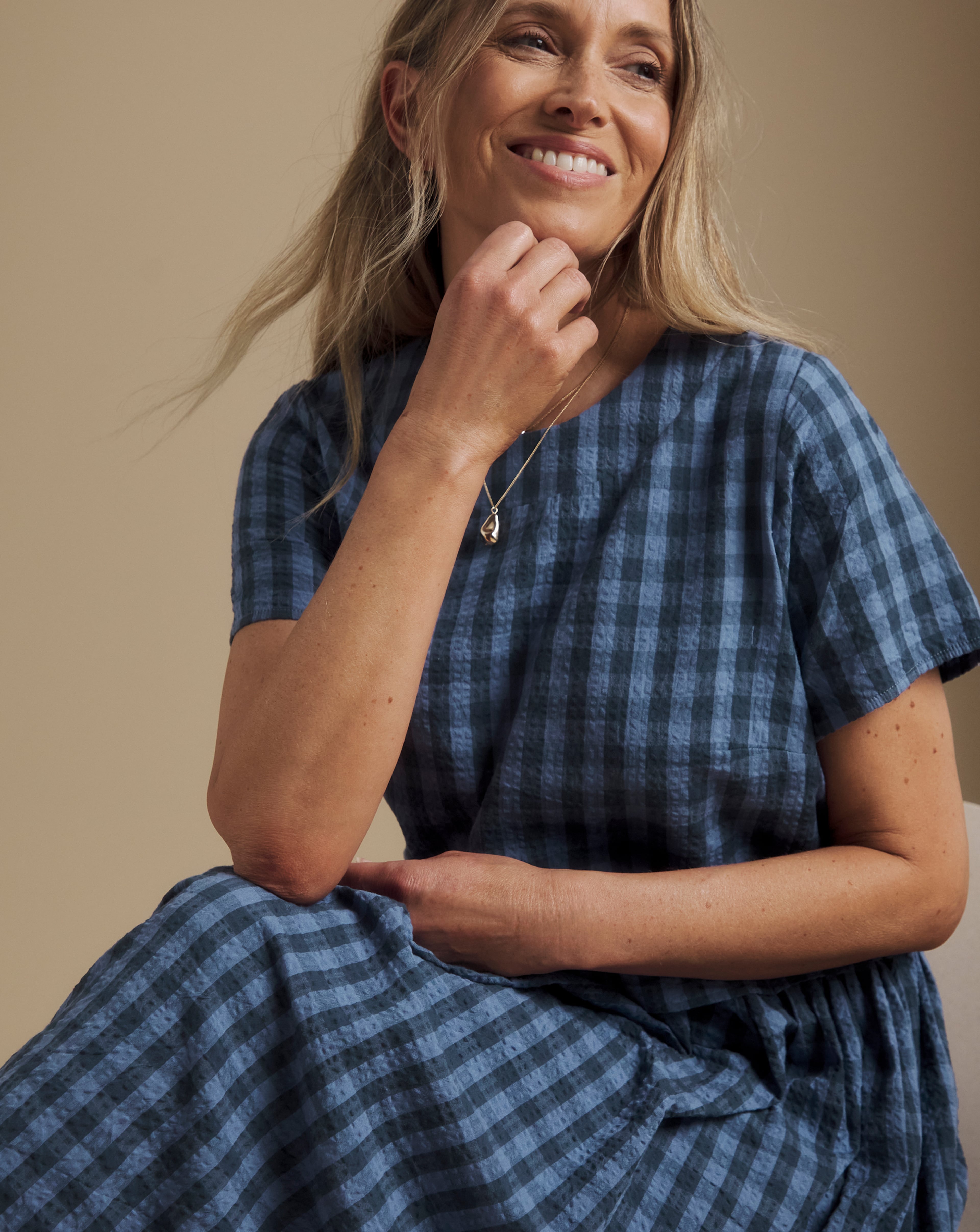 Close-up of a person seated wearing a Julipa blue check-pattern midi dress and pendant necklace.
