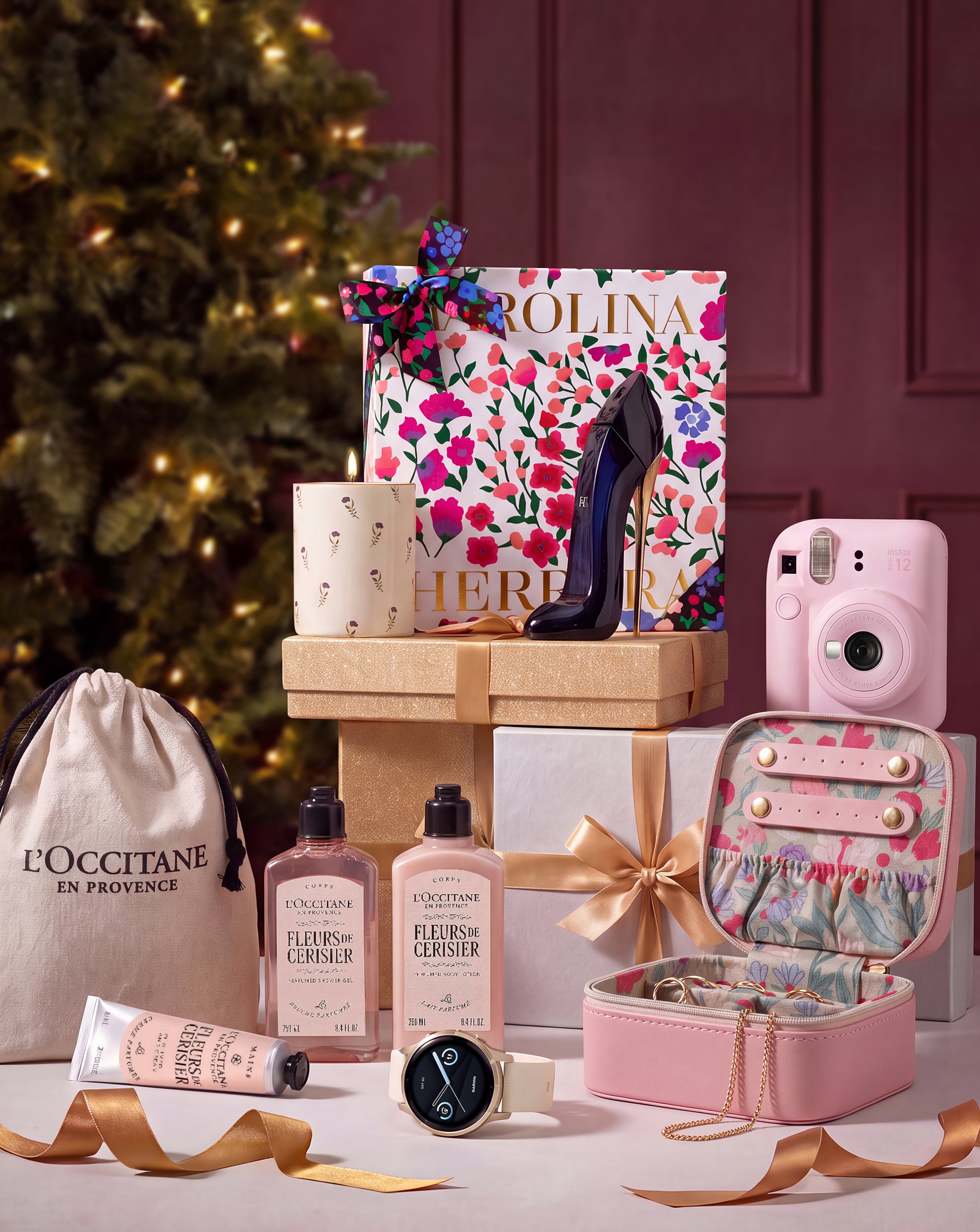 A Christmas gift arrangement featuring L'Occitane skincare products, Carolina Herrera perfume, a pink Instax camera, a watch, and a jewellery case.
