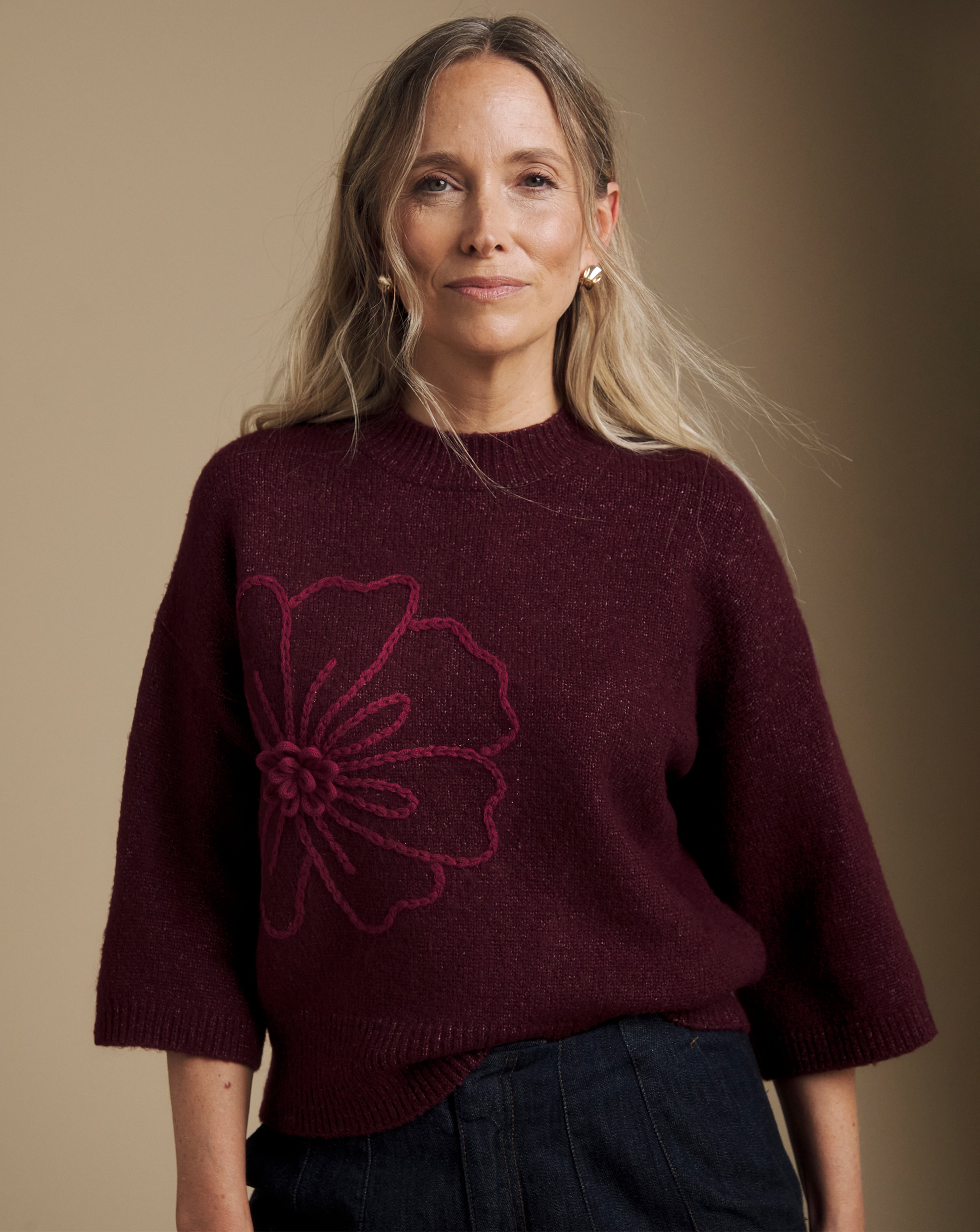 Person wearing a burgundy knit sweater with a large stitched flower design, paired with dark trousers.