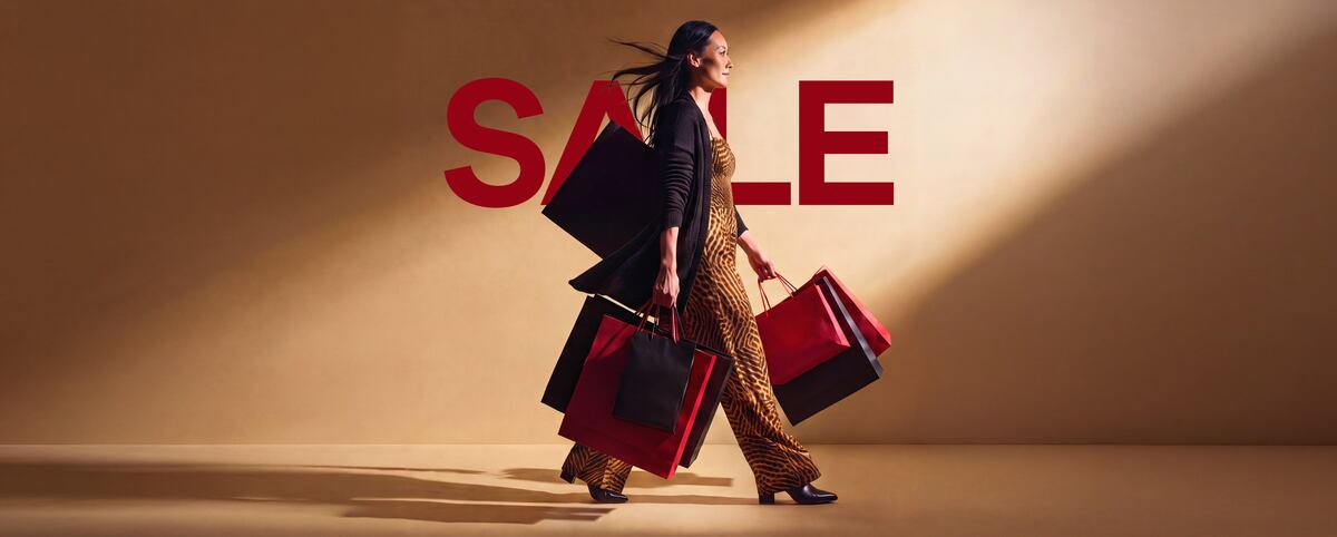A woman walking with several red and black shopping bags in front of a large red “Sale” sign.