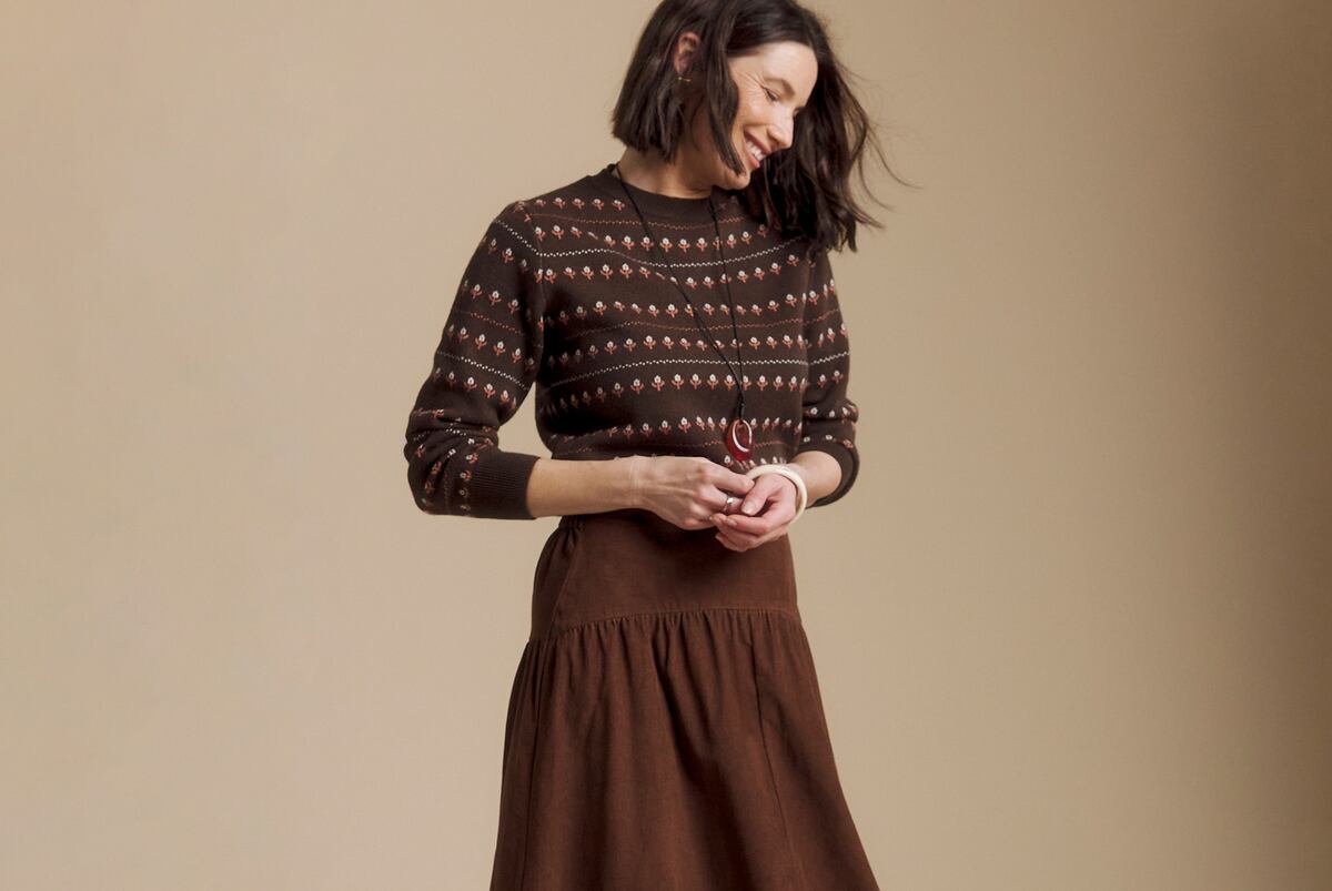 Person wearing a brown patterned knit sweater and flowing brown skirt, with a long pendant necklace.
