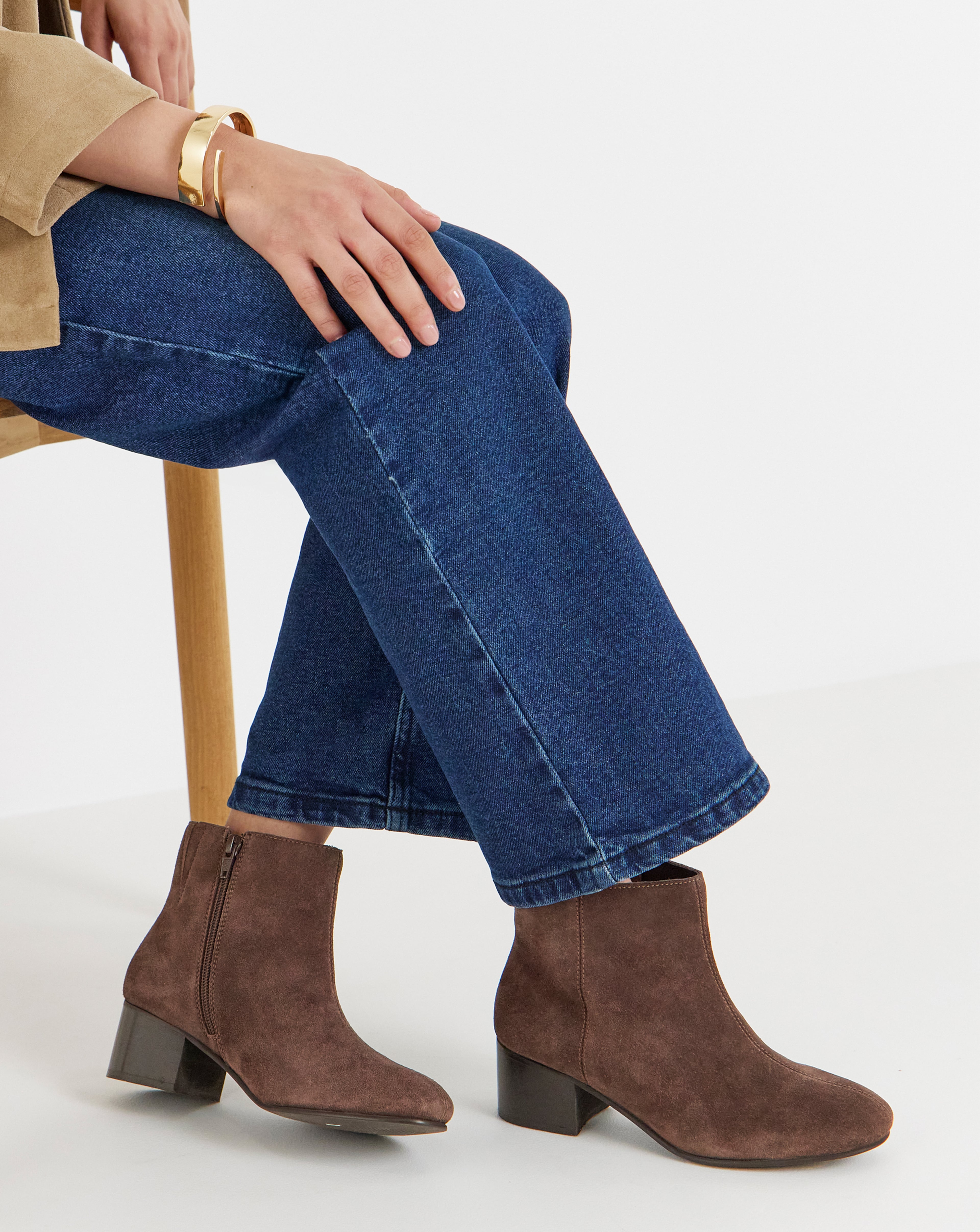 Close-up of a person seated wearing blue cropped jeans and brown suede ankle boots. 