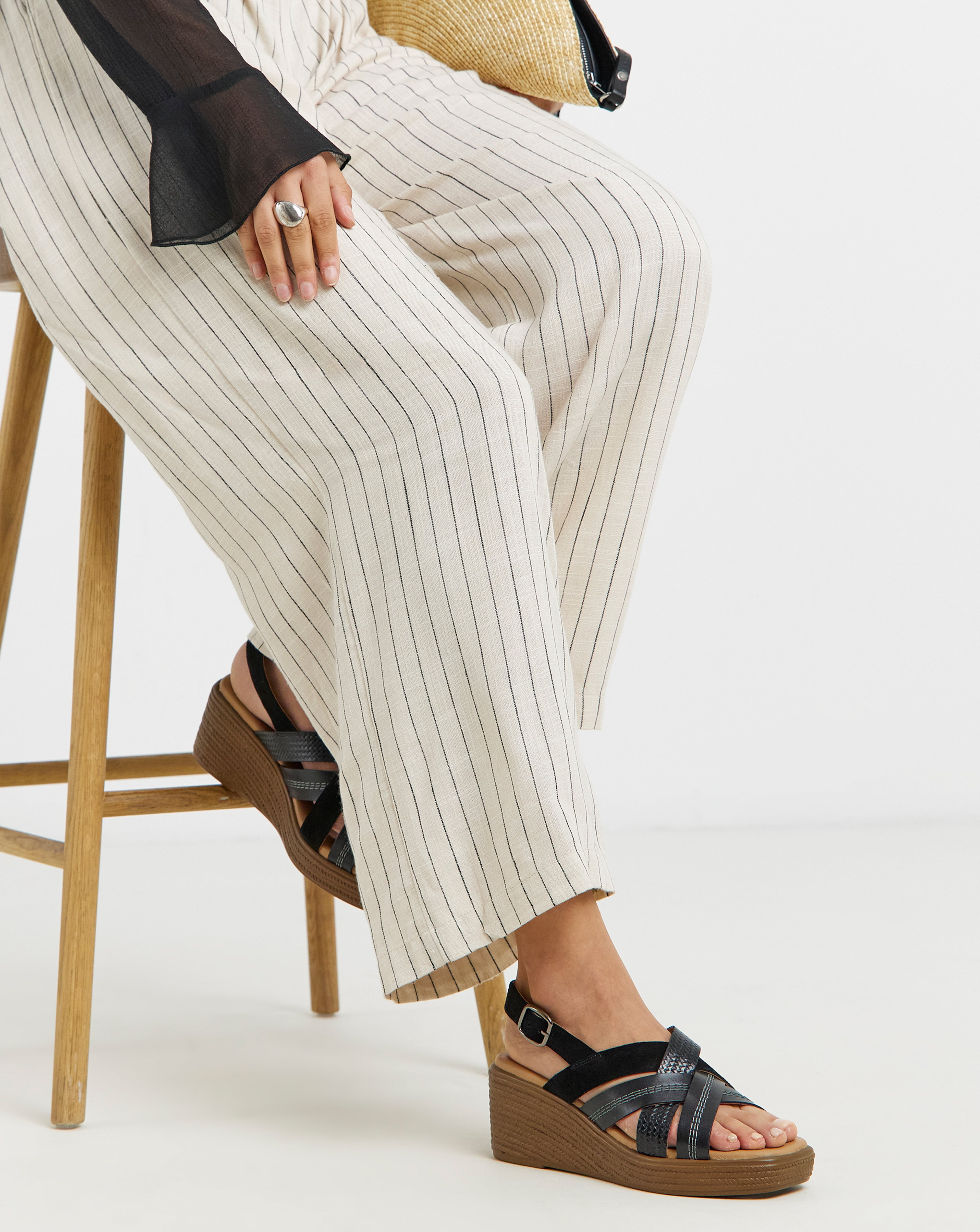 Close-up of a seated person wearing cream pinstriped trousers and black wedge sandals with straps.