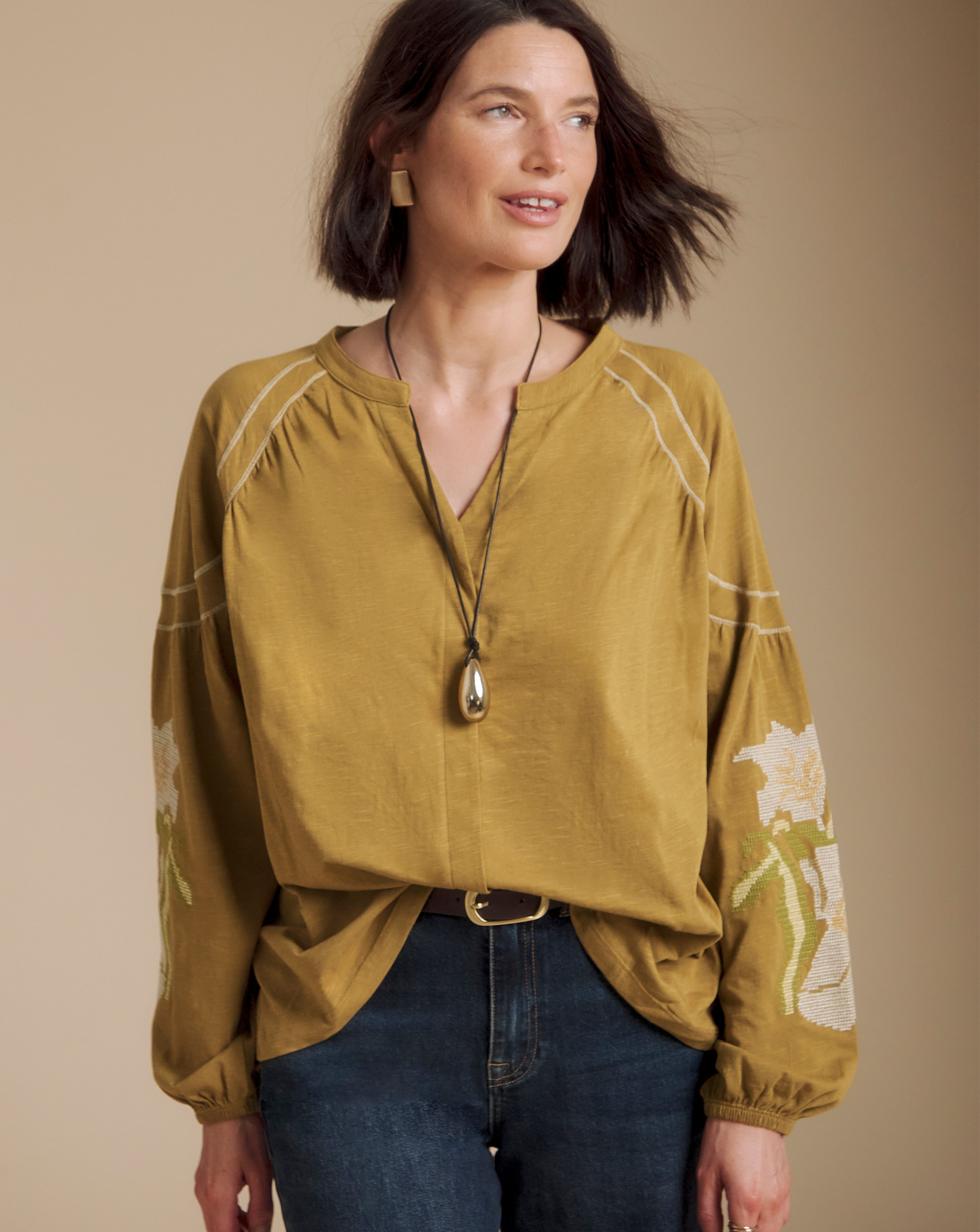 Person wearing a Julipa ochre-coloured blouse with embroidered sleeve details, dark blue jeans, and a pendant necklace in a studio setting.
