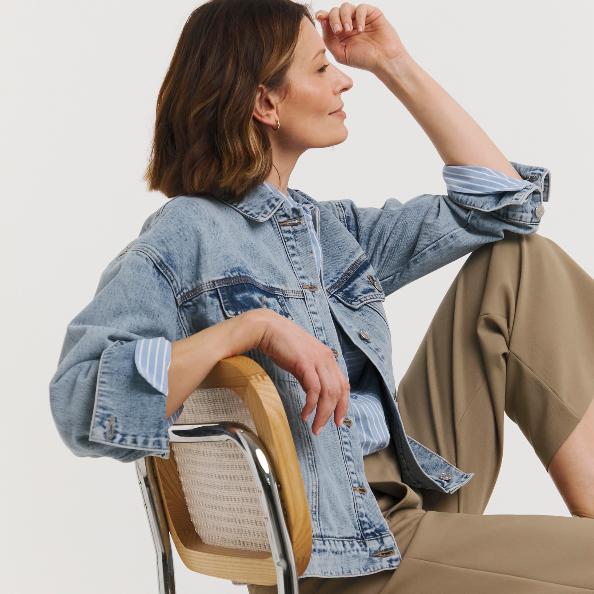 Person seated on a chair wearing a light-wash denim jacket layered over a blue striped shirt, paired with taupe cropped trousers.