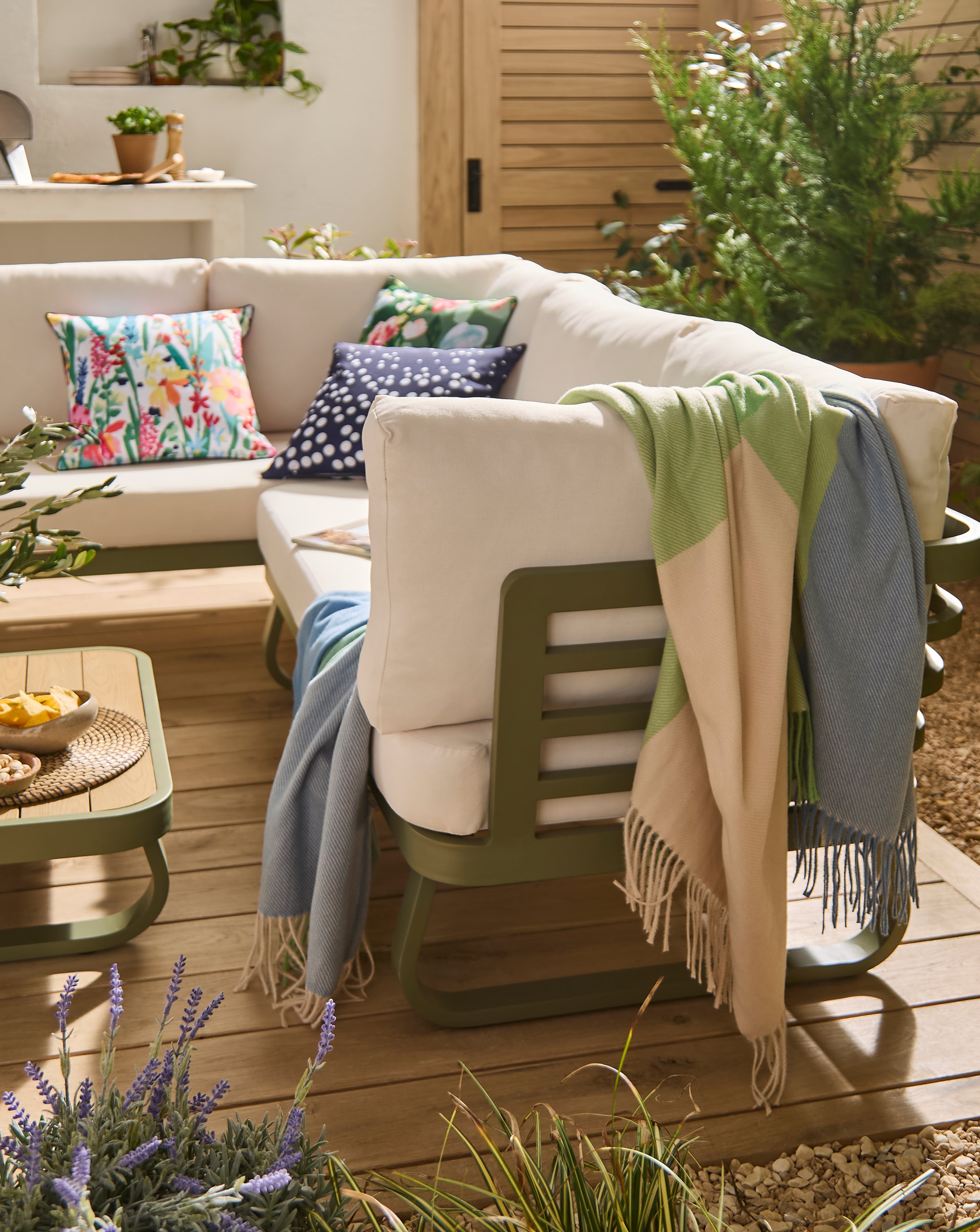 Outdoor seating area with a cushioned sofa, patterned cushions, and draped blankets on a wooden deck surrounded by plants.