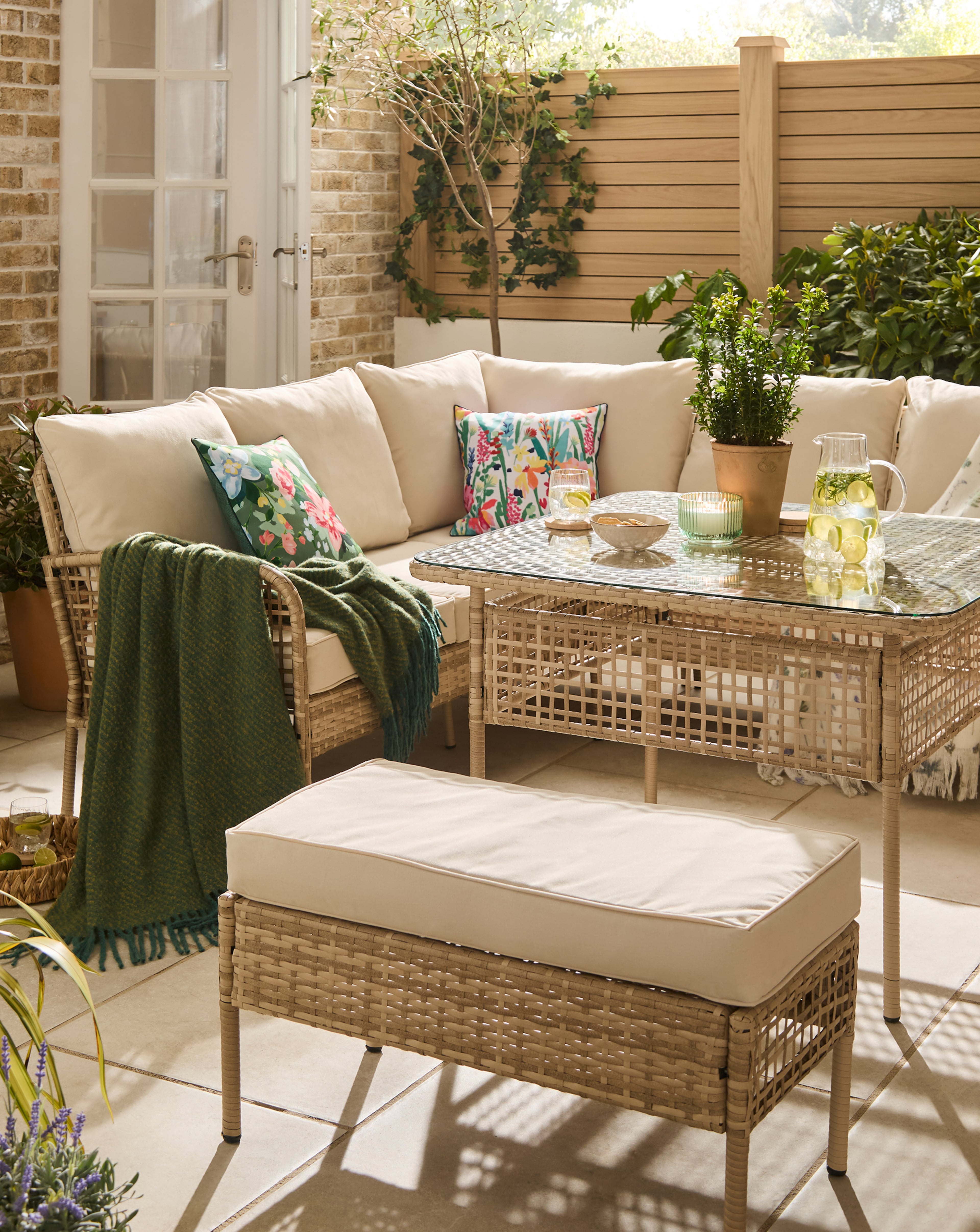 Outdoor patio seating area with a wicker sofa, cushioned bench, and glass‑top table, styled with patterned cushions.