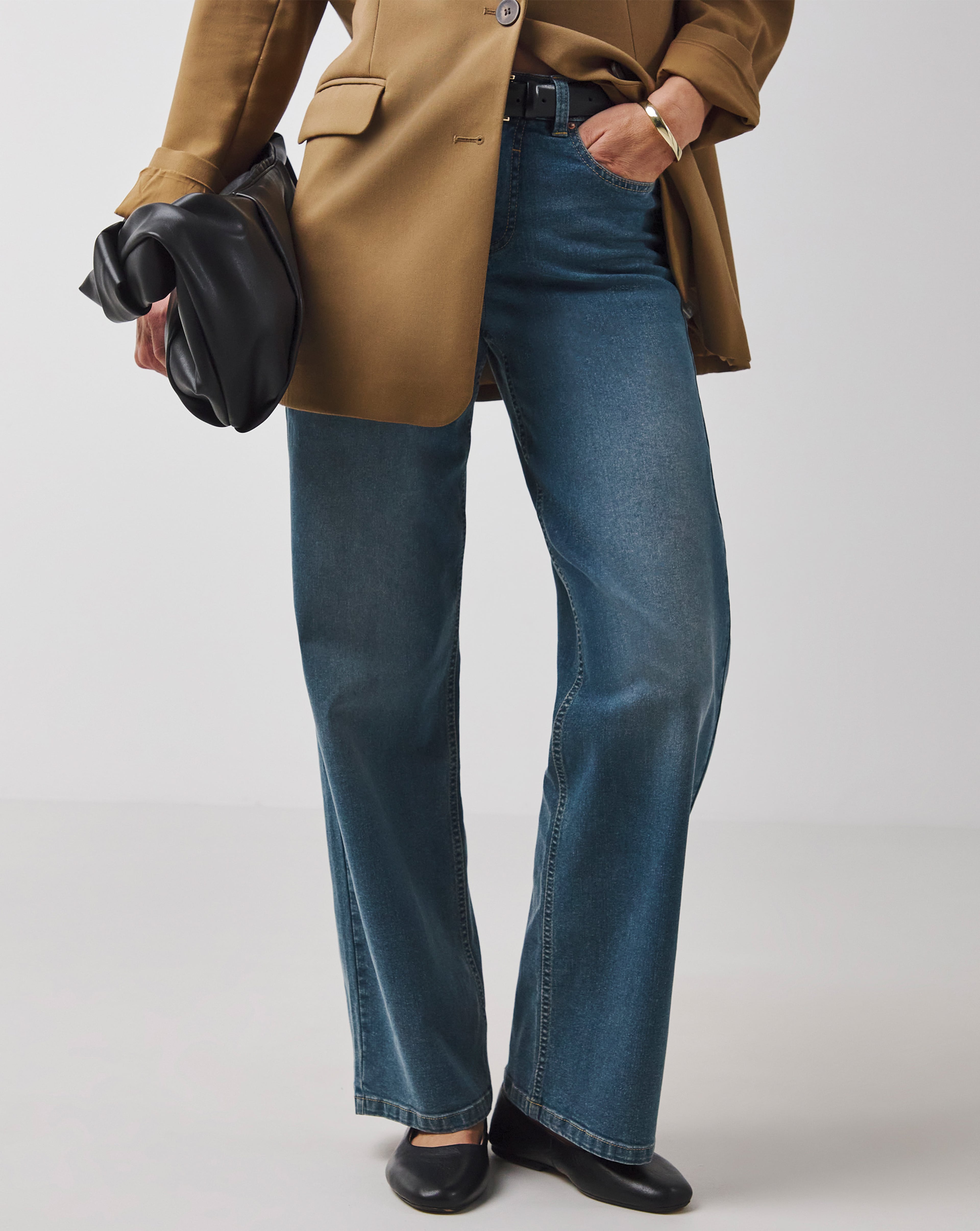 Person in a tan blazer, blue jeans, and black shoes holding a black handbag.