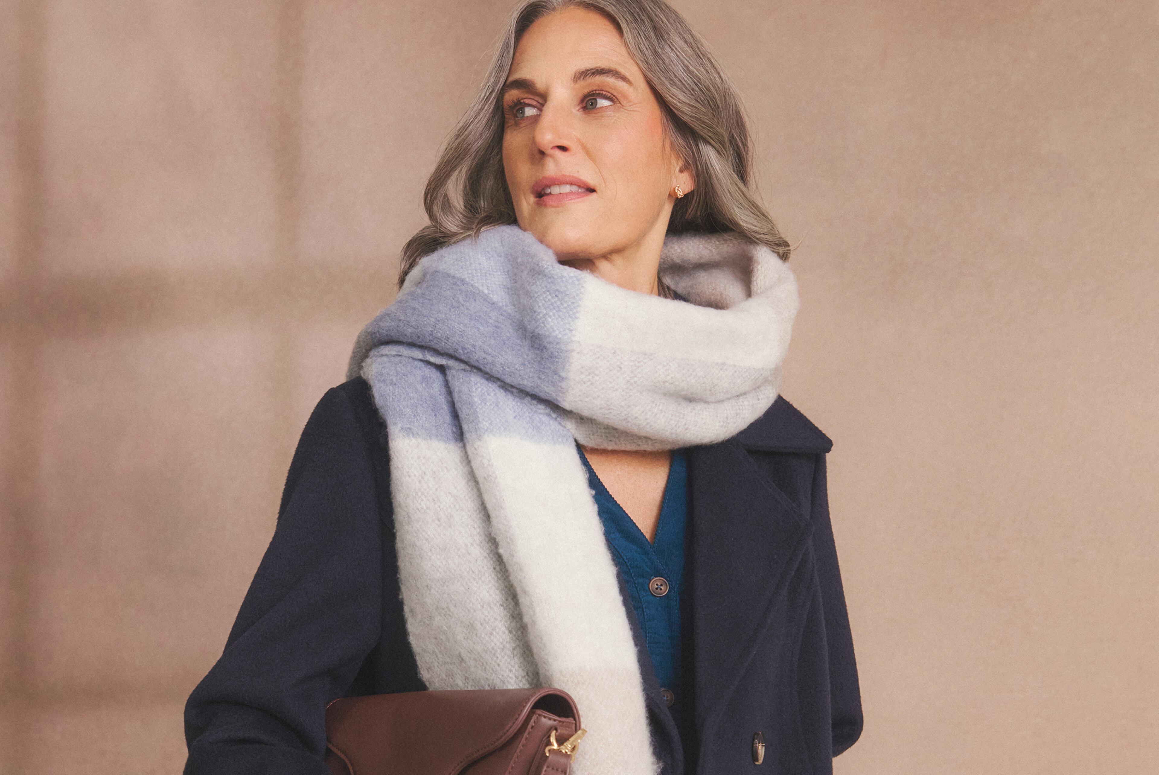 Person with long grey hair wearing a striped scarf and dark coat, holding a brown handbag