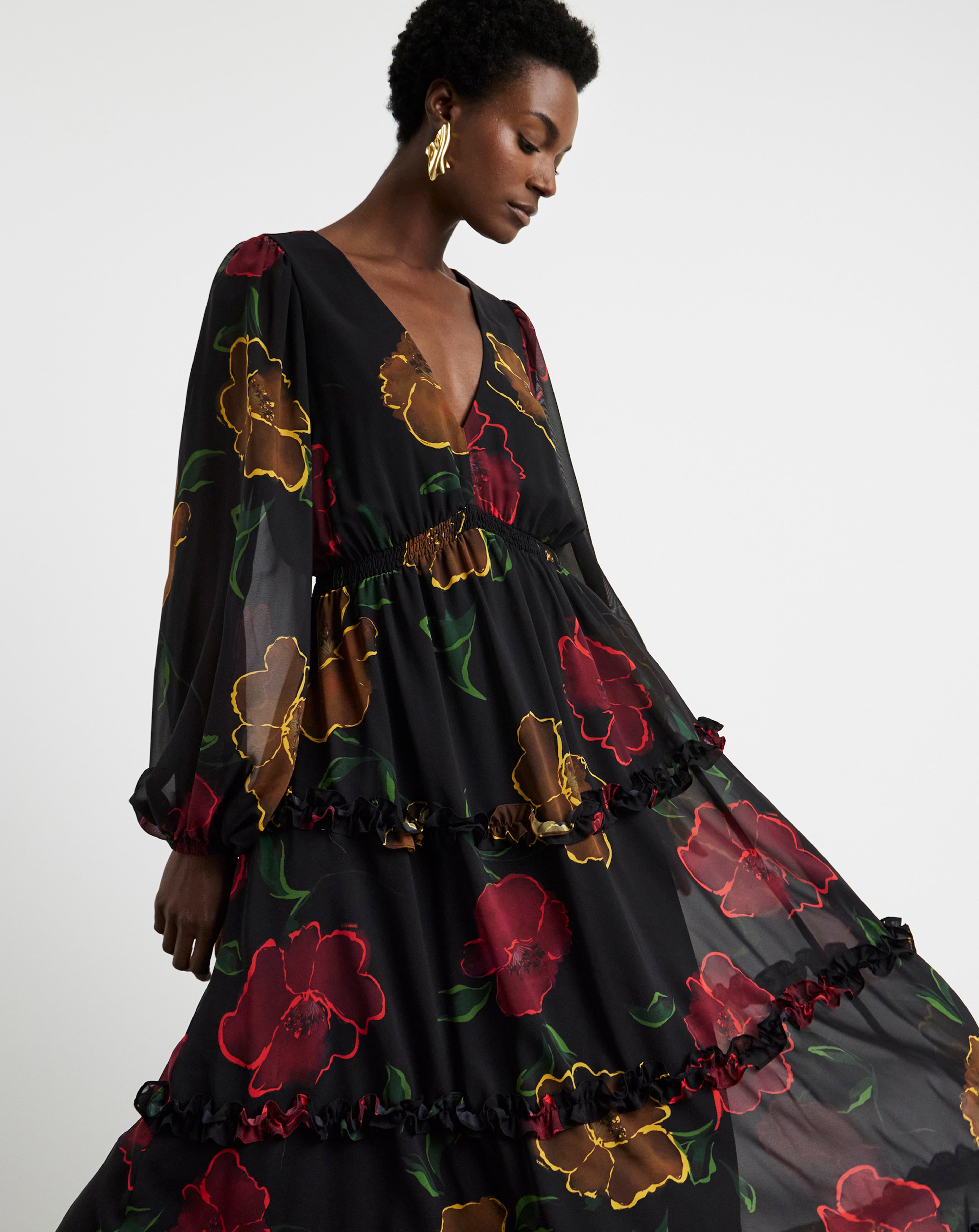Person in a long black dress with red and yellow floral patterns, sheer sleeves, and ruffled tiers.