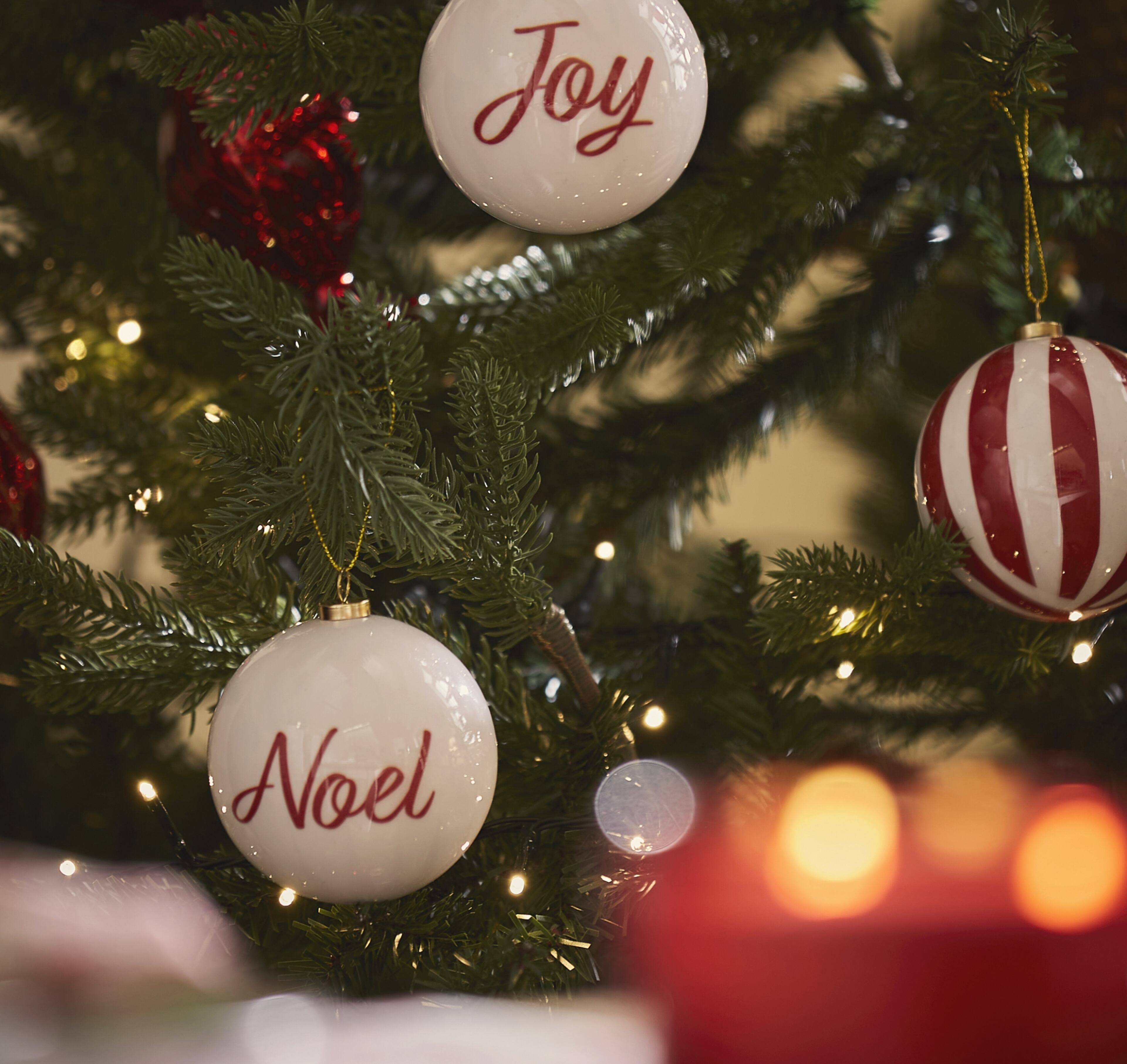 Close-up of classic Christmas ornaments hanging on a tree, including white baubles with "Joy" and "Noel" written in red.