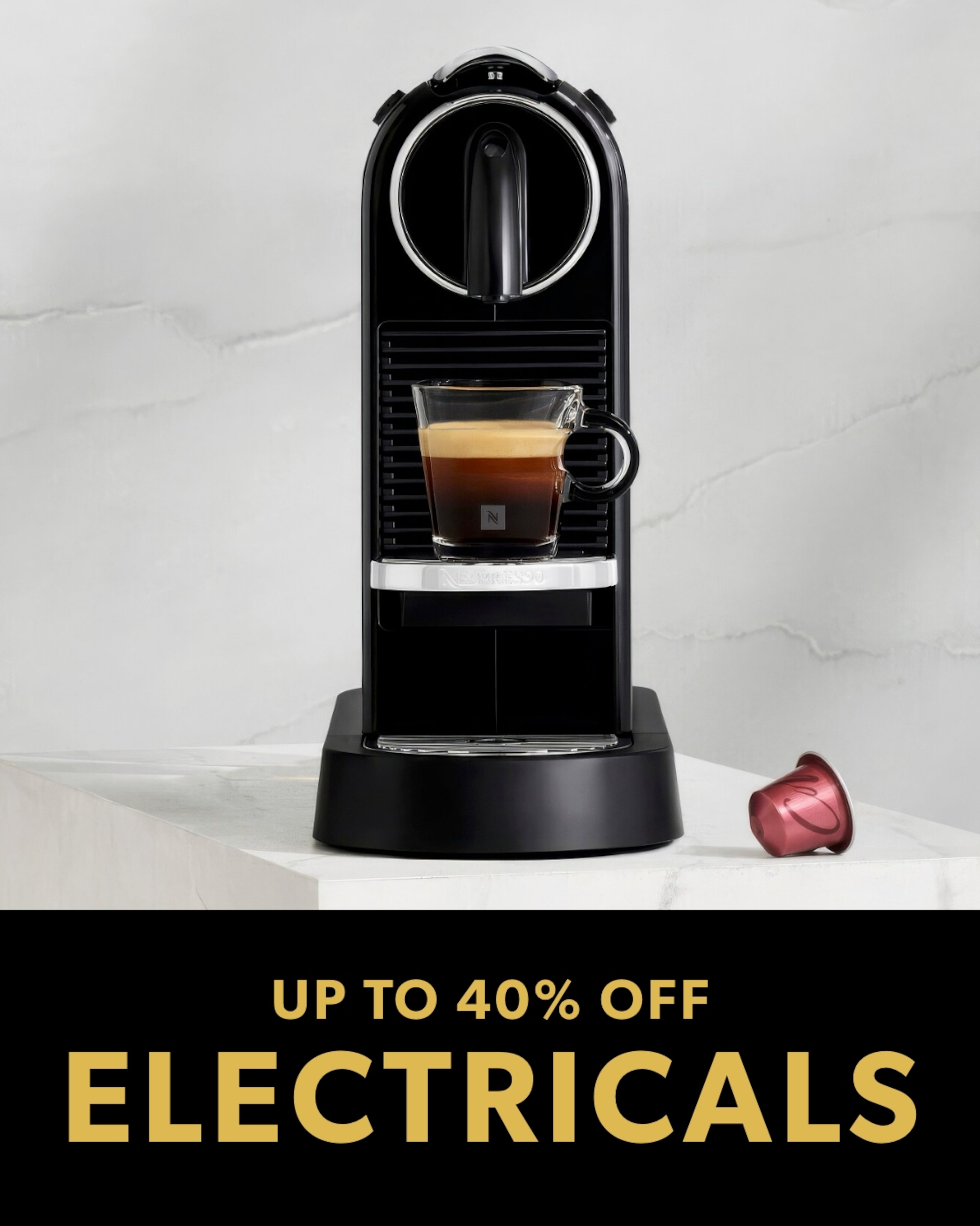 A black Nespresso-style coffee machine brewing espresso into a small glass cup on a marble counter, with the text "UP TO 40% OFF ELECTRICALS"