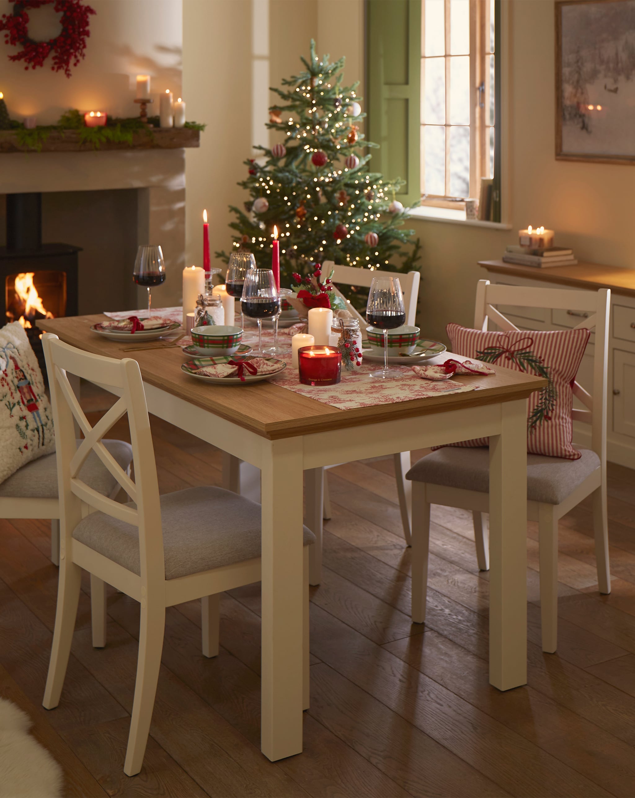 Dining room with a decorated table, Christmas tree, and fireplace with festive candles and a wreath.