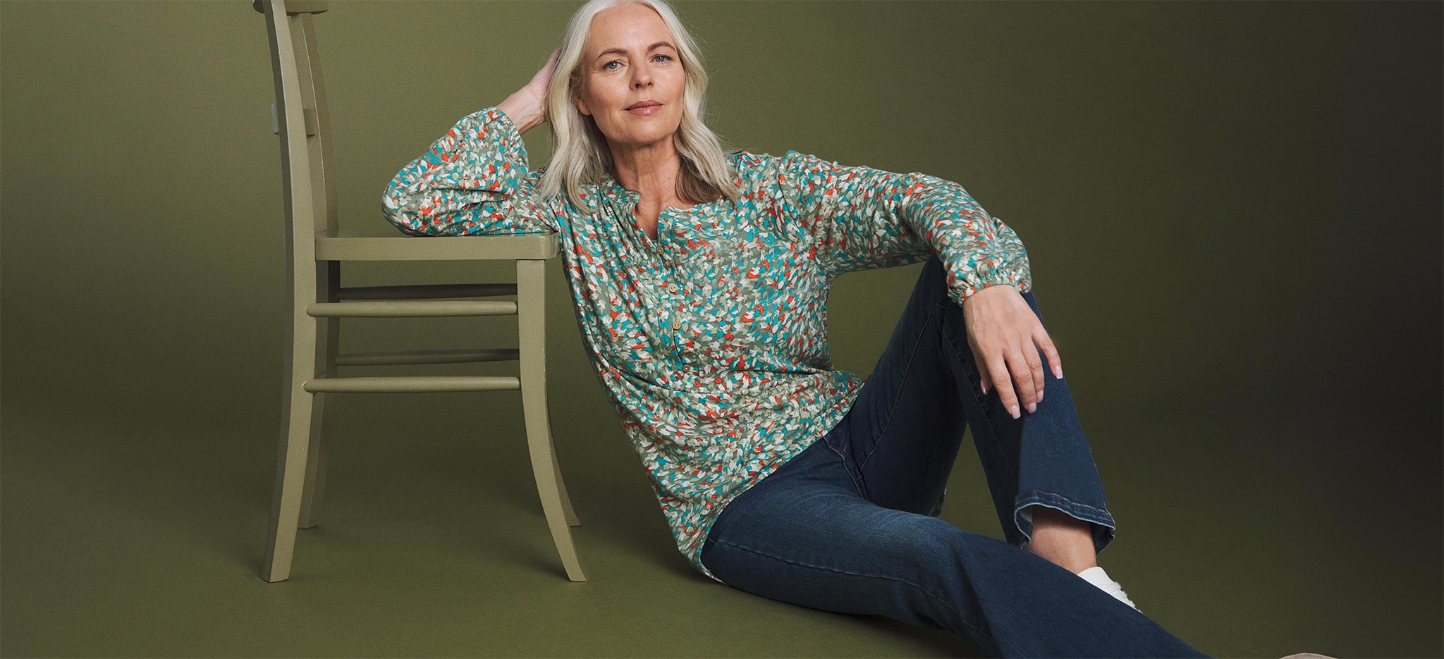 A woman sat on the flor wearing a green patterned blouse and blue jeans