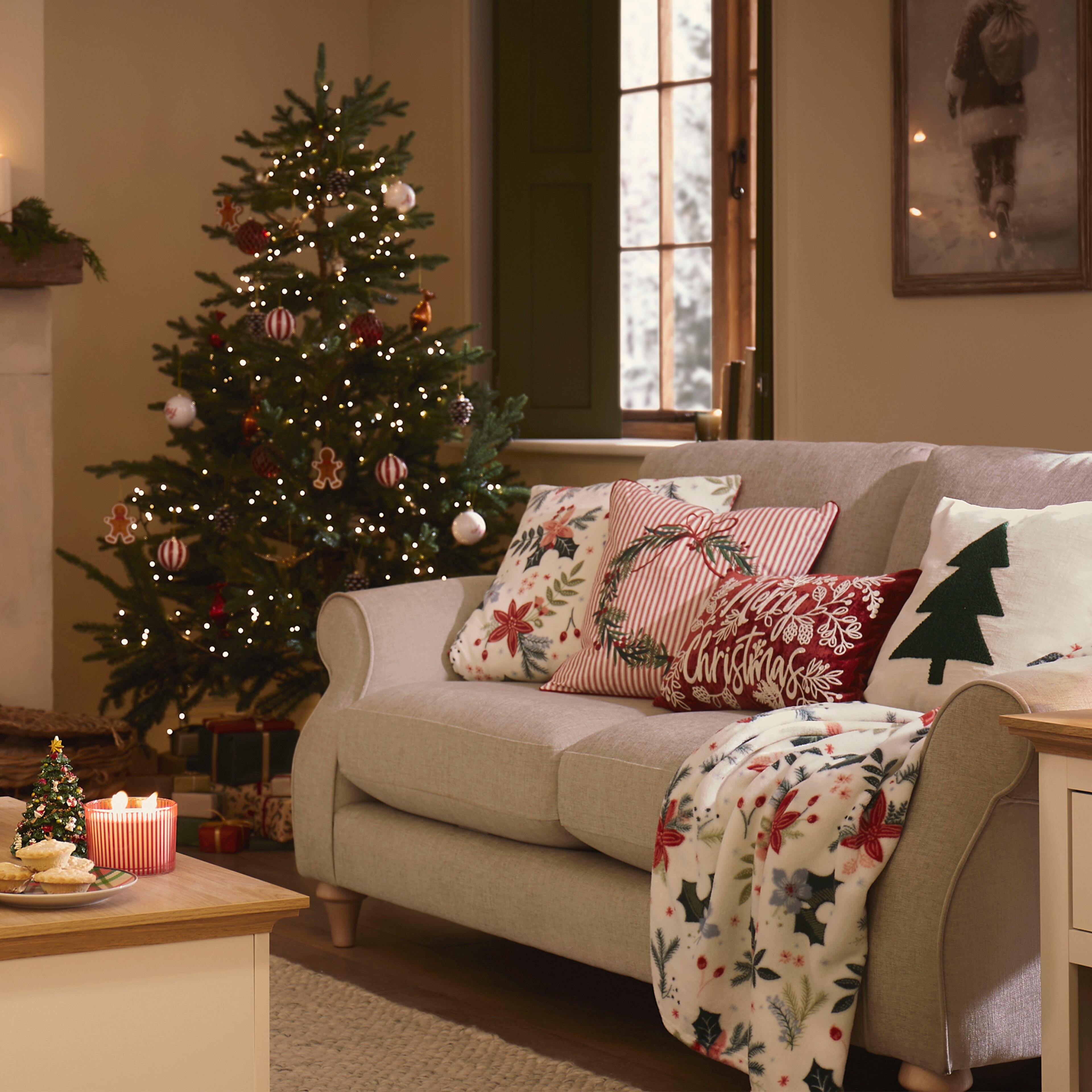 A living room with a decorated Christmas tree, a light grey sofa with festive cushions and throw, and a coffee table with candles and treats.