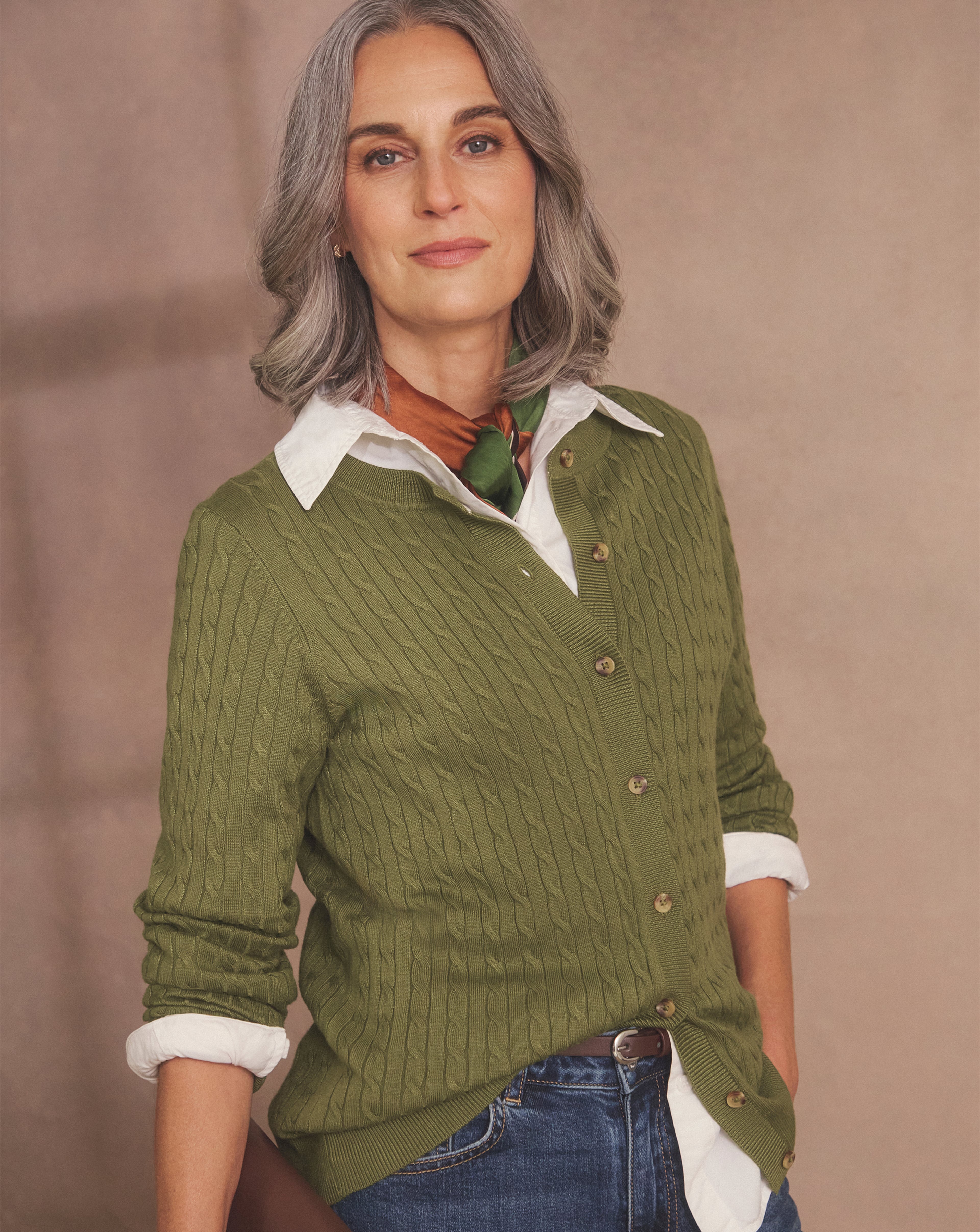 Person with grey hair wearing a green cable-knit cardigan, white shirt, scarf, and blue jeans.