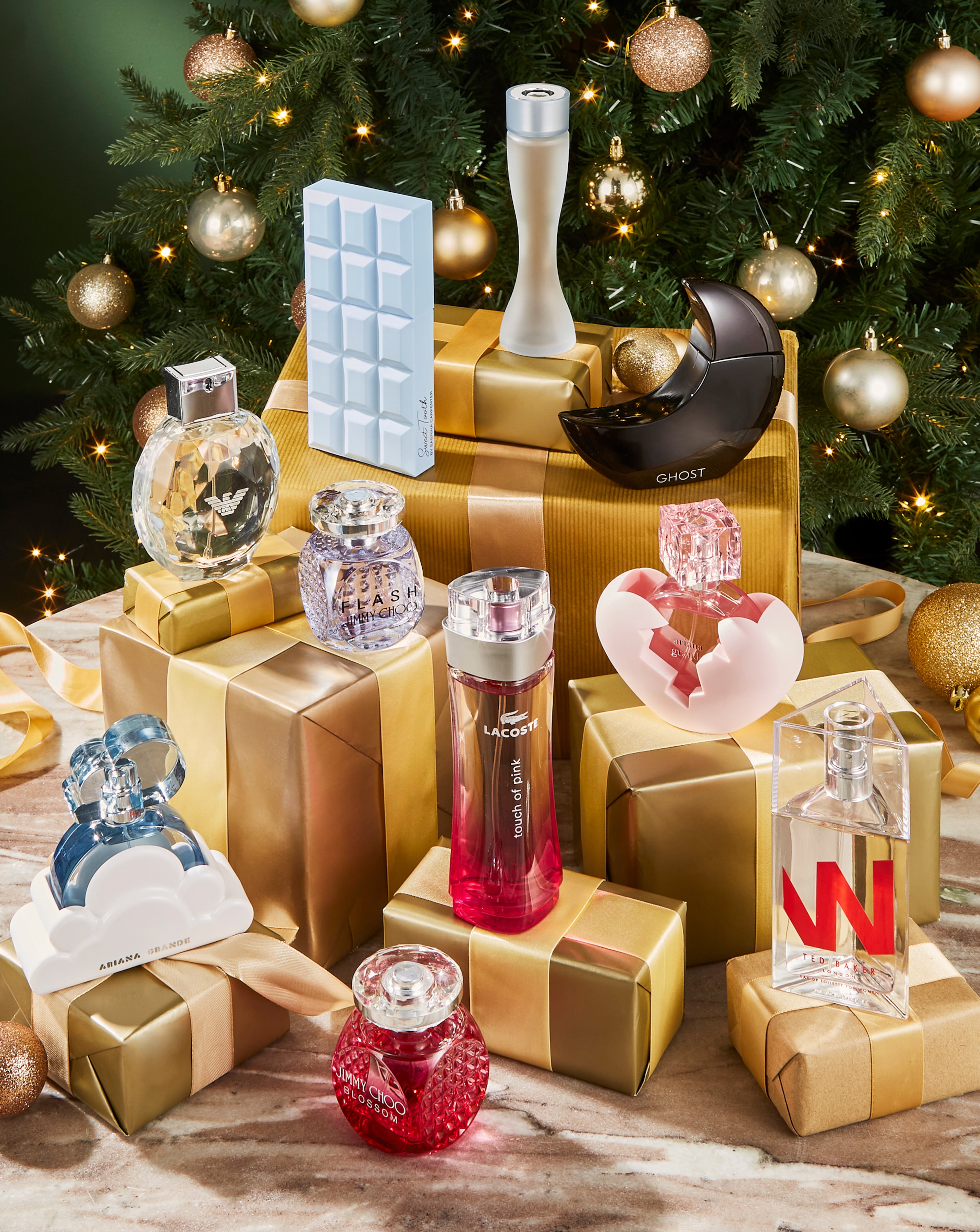 Assorted fragrance bottles arranged on gold-wrapped gifts in front of a decorated Christmas tree.